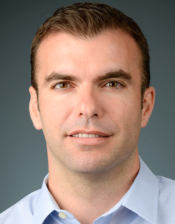 Headshot photograph of a man with short brown hair, slight smile, light blue shirt, gray background