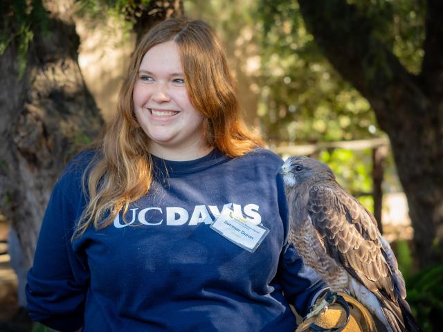Smiling young woman in UC Davis sweatshirt outdoors, hawk perched on gloved arm