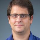 Headshot photograph of a man with glasses, blue shirt, neutral expression, gray background.